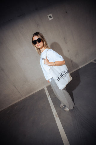 Woman holding a cream tote bag with collective logo in an indoor setting