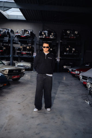 Person standing in a garage with cars on shelves in the background with a black quarter zip sweater.