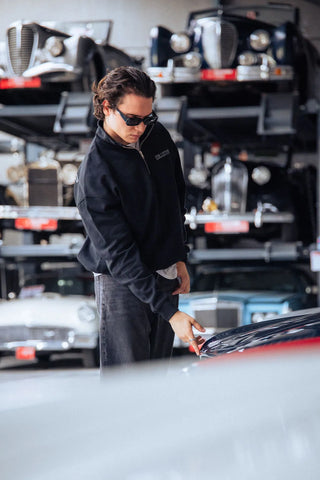 Person working on a car in a garage with vintage cars in the background while wearing a black quarter zip sweater.