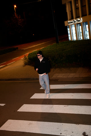 Person crossing a street at night with a building in the background. Wearing a black quarter zip sweater.