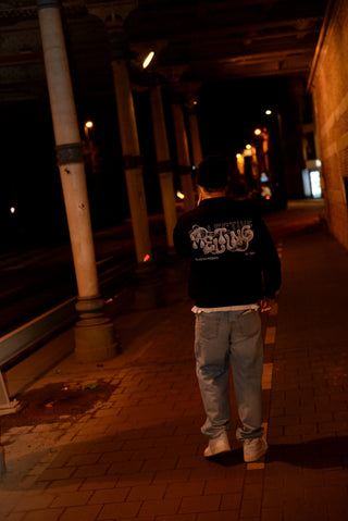 Person walking away on a dimly lit street at night. He's wearing a black quarter zip hoodie with white letters.