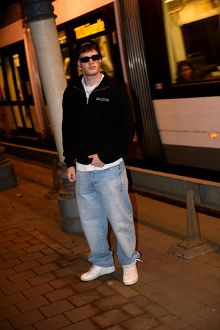 Person wearing a black quarter zip sweater and sunglasses standing on a sidewalk with a tram in the background.