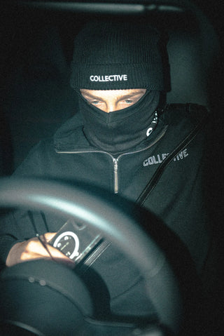 Person wearing a black quarter zip sweater and beanie with 'Collective' branding, sitting inside a vehicle.