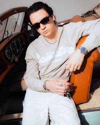 Person wearing sunglasses and a beige sweatshirt sitting inside a vintage aston martin.