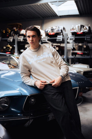 Person leaning against a classic car in a garage setting wearing a beige sweater